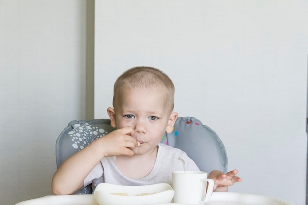 Un petit garçon avec une coupe de cheveux courte mange indépendamment et avec appétit de la nourriture et boit du jus d'un tube dans une chaise d'enfant