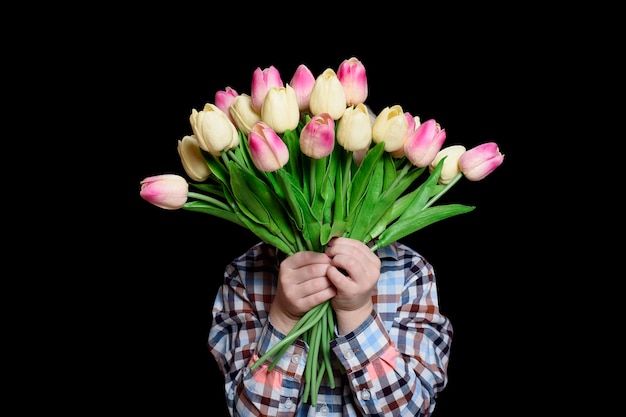 Petit garçon couvre son visage avec un bouquet de tulipes. Isoler sur fond noir.