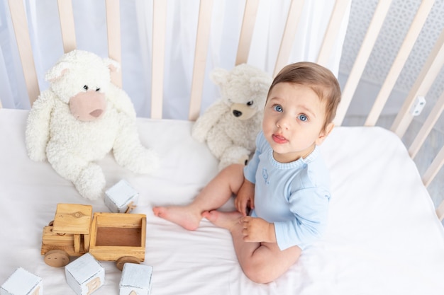 Petit garçon dans le berceau dans la chambre des enfants avec des jouets en bois sur un lit en coton blanc jouant