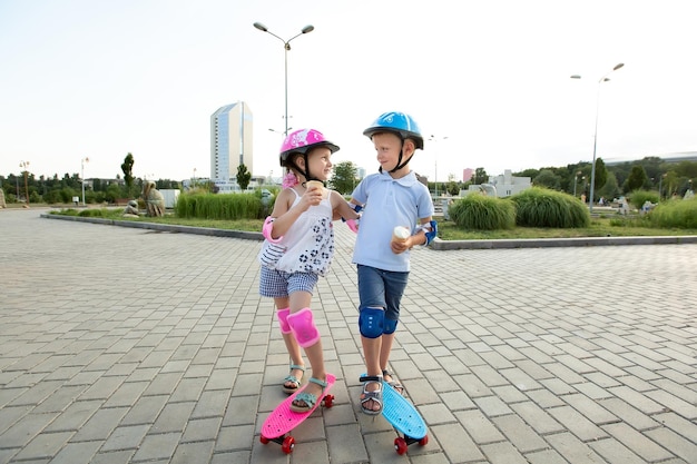 Un petit garçon dans un casque embrasse une fille dans le parc, ils font du skateboard et mangent de la crème glacée