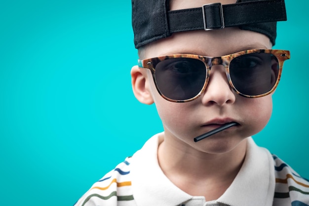 Petit garçon dans une casquette de baseball et des lunettes de soleil sur un fond turquoise garçon cool avec une sucette sur sa joue