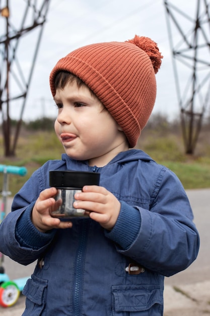 Un petit garçon dans une casquette orange dans la nature boit du thé chaud dans un thermos