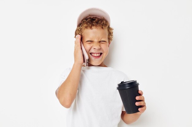 Petit garçon dans une casquette de t-shirt blanc avec un téléphone dans un verre avec une boisson fond clair inchangé