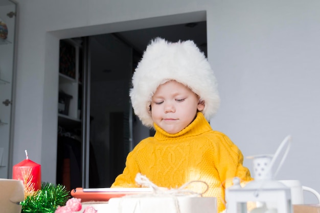 Un petit garçon dans un chandail jaune et un chapeau de Père Noël rouge à une table en bois avec une tablette et dans des décorations de Noël