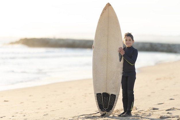 Un petit garçon dans une combinaison tenant une grande planche de surf blanche debout sur le bord de la mer