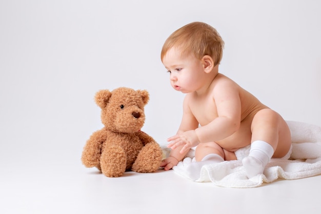 Le petit garçon dans une couche est assis sur un fond blanc avec un ours en peluche