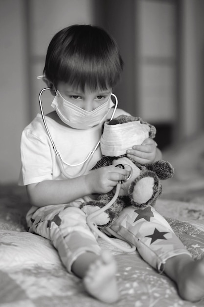 Un petit garçon dans un masque assis sur le lit et stétoscope un ours en peluche Image avec mise au point sélective