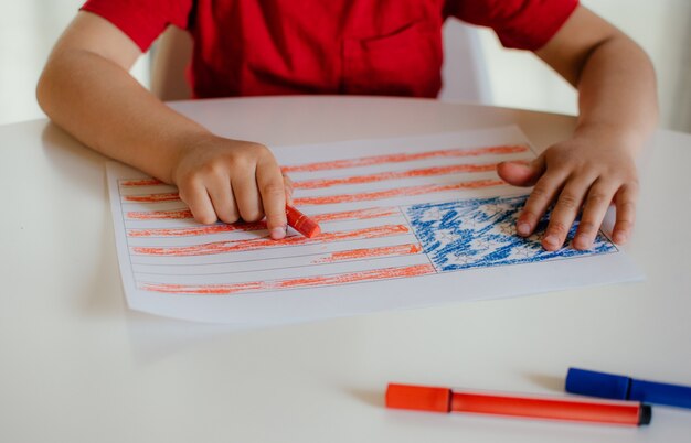 Un petit garçon dans un t-shirt rouge peint le drapeau américain sur un tableau blanc. Dessin pour enfants du drapeau de l'Amérique. Fête de l'indépendance, 4 juillet.