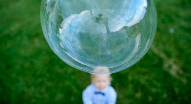 Photo petit garçon debout sur l'herbe floue regarde de bas en haut la grosse bulle avec un reflet. photo de haute qualité