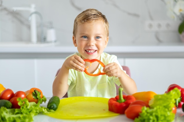 Petit garçon enfant assis à la table avec des légumes dans la cuisine à la maison manger des légumes bébé souriant concept de saine alimentation végétarien