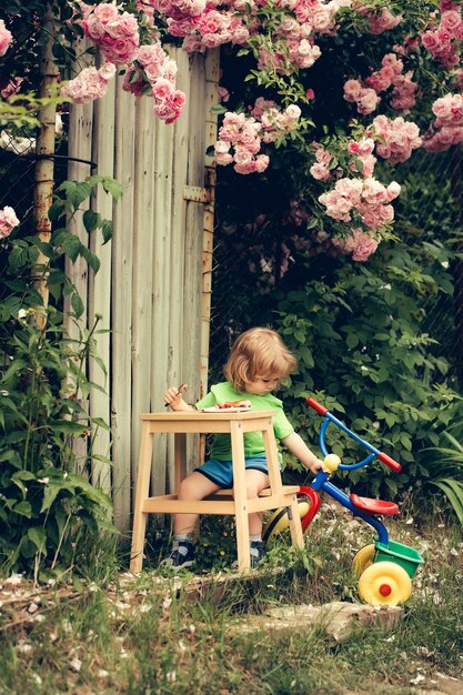 Petit garçon enfant aux longs cheveux blonds assis à une table en bois mangeant une tarte près d'un jouet en plastique en plein air avec...