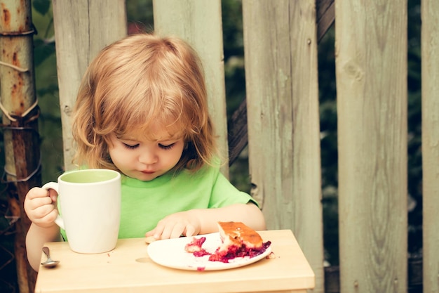 Petit garçon enfant aux longs cheveux blonds et au visage sérieux assis à une table en bois mangeant une tarte aux baies et un biscuit avec une tasse de lait en plein air près d'une clôture en bois