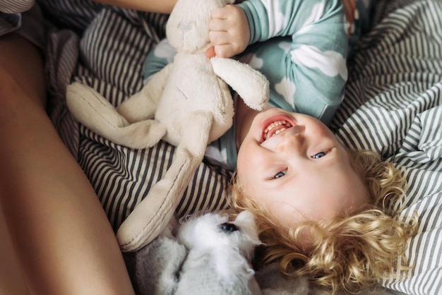 Petit garçon enfant bouclé aux cheveux blonds en pyjama bleu joue des rires se trouve sur le lit