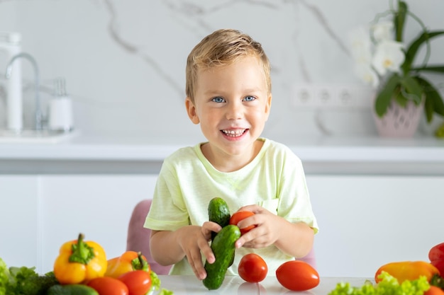 Un petit garçon enfant est assis à une table avec des légumes dans la cuisine à la maison tenant du poivre dans ses mains le bébé sourit le concept d'une alimentation saine végétarienne