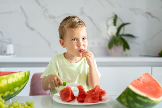 Petit garçon enfant est assis à la table avec des pastèques dans la cuisine de la maison il y a une pastèque le bébé sourit le concept d'une alimentation saine végétarienne