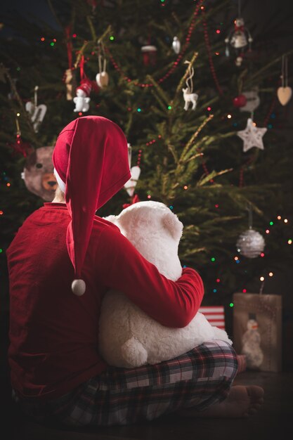 Un petit garçon est assis à côté d'un sapin à feuilles persistantes décoré de guirlandes et d'autres jouets du Nouvel An