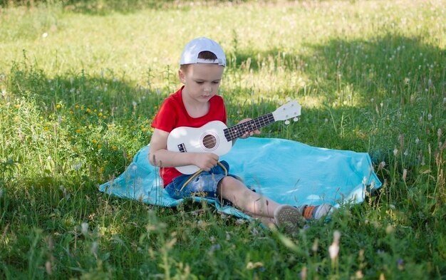 Un petit garçon est assis sur l'herbe dans le parc en été et joue du ukulélé Happy moments guitar closeup baby Un enfant charmant et joyeux profite d'un jeu de plein air d'été sain