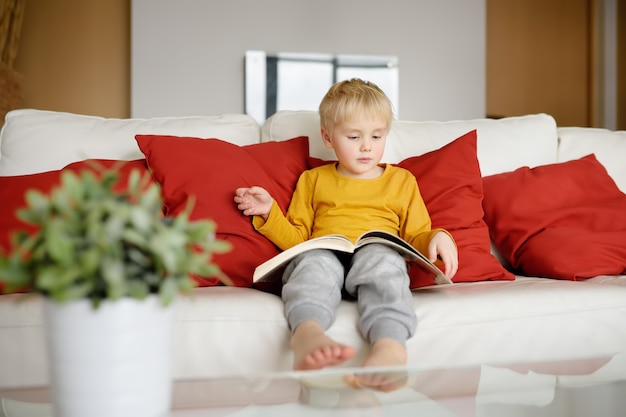 Petit garçon est assis à la maison sur le canapé et lit un livre. Apprendre à lire.