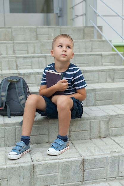 Un petit garçon est assis sur les marches de l'école, tenant des cahiers et regardant le ciel