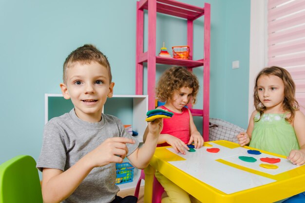 Le petit garçon fait la démonstration de son produit en pâte à modeler Les enfants sont assis au bureau dans la salle de jeux