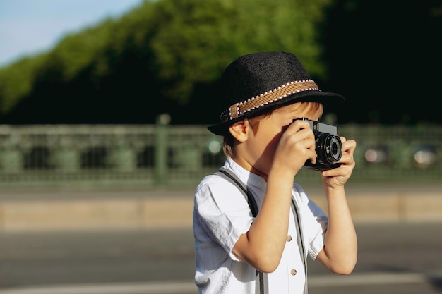 Un Petit Garçon Fait Semblant De Prendre Une Photo Avec Un Appareil Photo Rétro, Il Se Tient Sur Le Trottoir D'un Pont En Ville