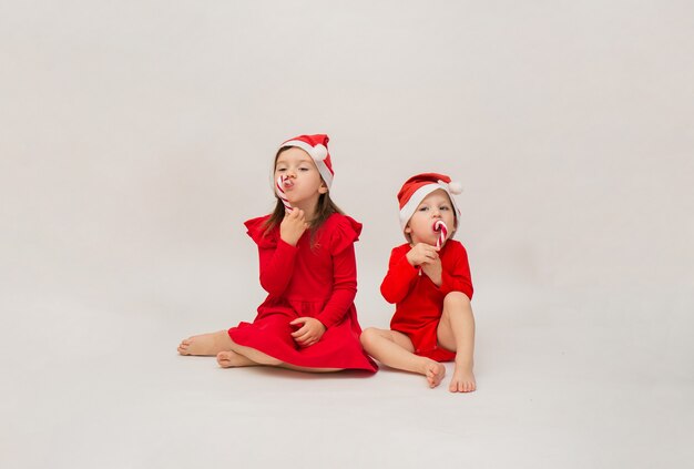 Petit garçon et fille en bonnets rouges s'asseoir avec des sucettes sur blanc