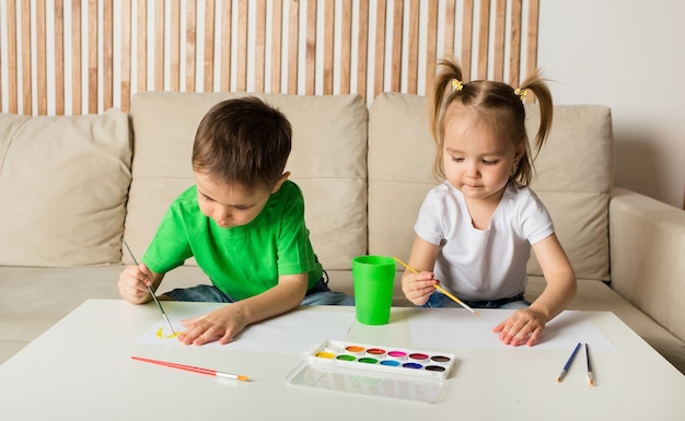 Petit garçon et une fille dessiner avec un pinceau et peindre sur papier à une table dans une pièce