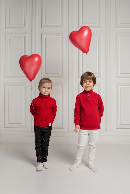 Un petit garçon et une fille tiennent des ballons en forme de coeur et regardent la caméra sur un fond blanc