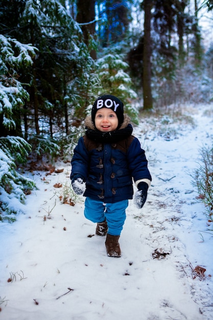 Un petit garçon gai de 2 ans dans une veste bleue se promène dans la forêt d'hiver