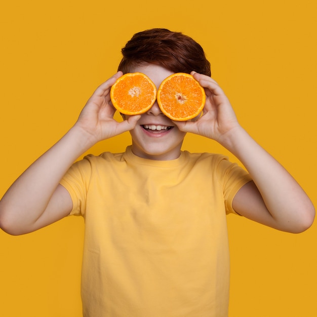 Petit garçon de gingembre caucasien se couvre les yeux d'oranges posant dans un t-shirt sur un mur jaune avec un espace libre