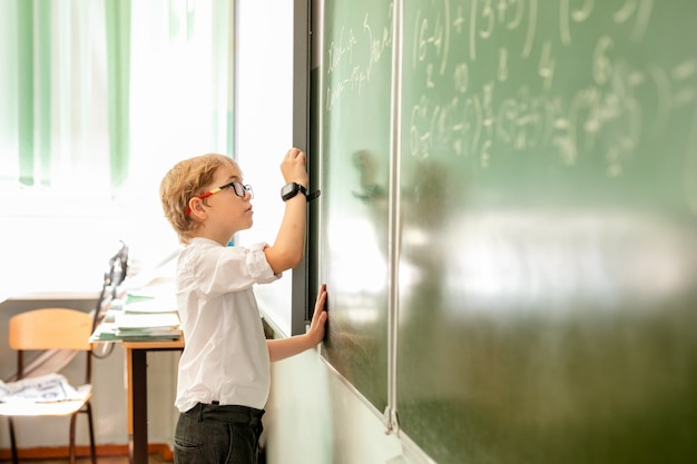 Petit garçon avec de grandes lunettes noires et une chemise blanche se tenant près du tableau de l'école avec un morceau de craie faisant face intelligente