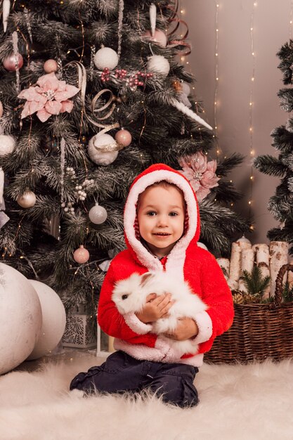 Petit garçon habillé en père noël tenant un lapin dans ses mains près de l'arbre de noël