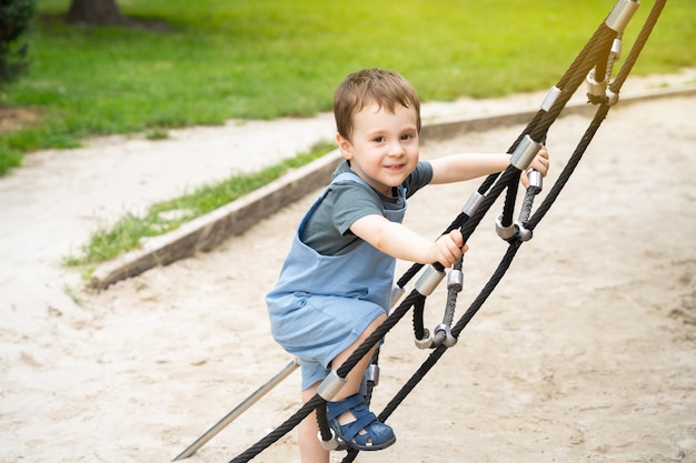 Un petit garçon heureux de 25 ans grimpe sur l'échelle à corde dans l'aire de jeux du parc. Jeux et divertissements actifs pour les enfants un jour d'été ensoleillé.