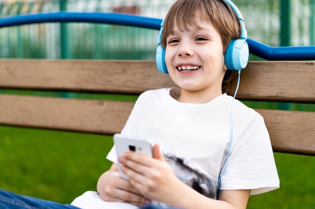 Un petit garçon heureux dans la rue est assis sur un banc avec des écouteurs et un téléphone et sourit