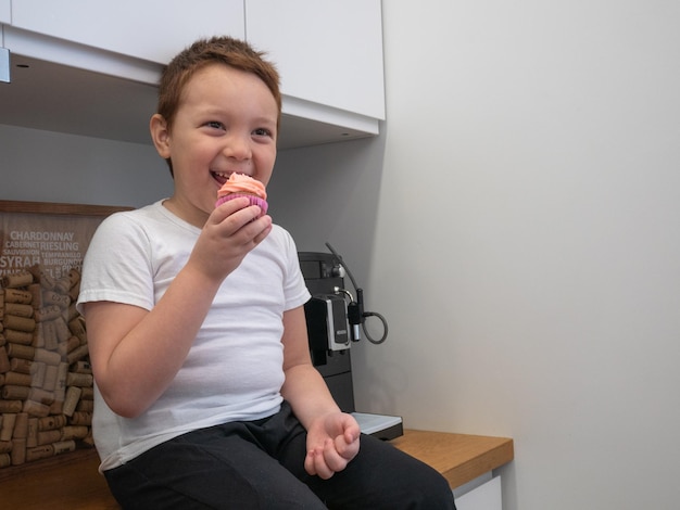 Un petit garçon heureux est assis sur la table de la cuisine en train de manger un petit gâteau en souriant