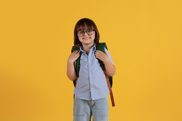 Petit garçon intelligent enfant d'âge préscolaire portant des lunettes souriant à la caméra avec sac à dos fond jaune