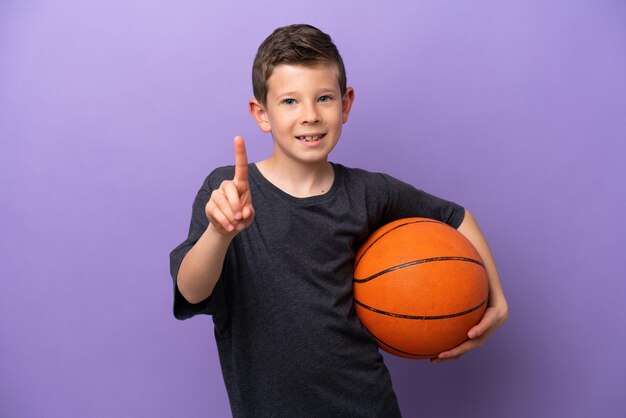 Petit garçon jouant au basket-ball isolé sur fond violet montrant et levant un doigt