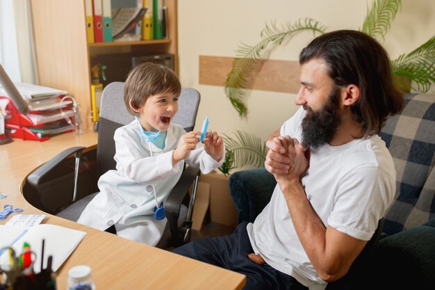 Un petit garçon jouant fait semblant d'être un médecin examinant un homme dans un cabinet médical confortable. Concept de soins de santé, d'enfance, de médecine, de protection et de bonheur. S'amuser, rire en donnant des pilules