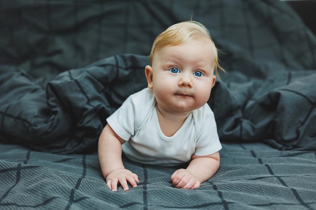 Un petit garçon joyeux avec de beaux grands yeux est allongé sur le lit à la maison dans un maillot de bain blanc sur une literie grise l'enfant est étendu sur le lit chez lui