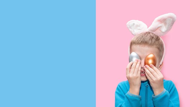 Un petit garçon joyeux dans une veste bleue et avec des oreilles de lapin tient des oeufs de Pâques colorés sur un fond bleu rose