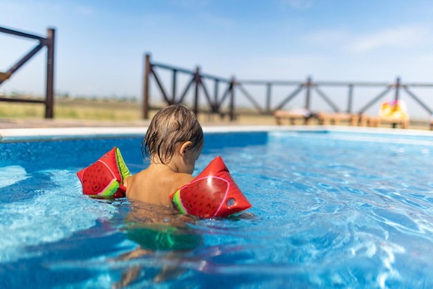 Un petit garçon joyeux et joyeux dans des brassards gonflables lumineux nage dans une piscine bleue pour les enfants avec de l'eau claire et transparente sous le chaud soleil d'été