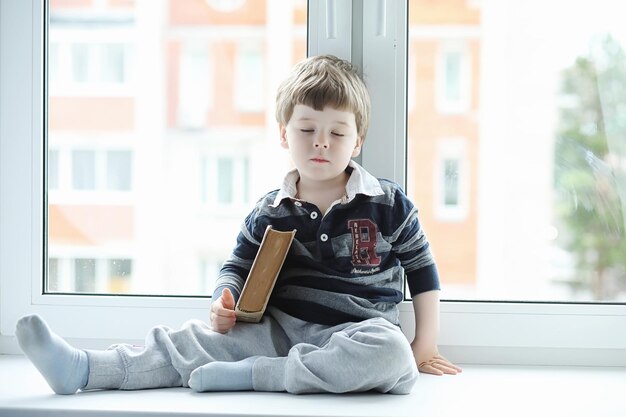 Le petit garçon lit un livre L'enfant est assis à la fenêtre et se prépare pour les cours Un garçon avec un livre dans les mains est assis sur le rebord de la fenêtre