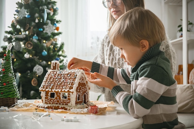 Petit garçon avec maman décorant la maison de pain d'épice de Noël ensemble, activités familiales et traditions à Noël et au Nouvel An