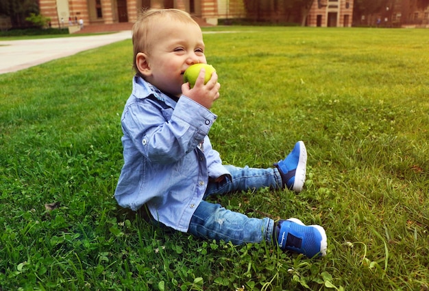 Petit garçon mange une pomme verte assis sur l'herbe et souriant