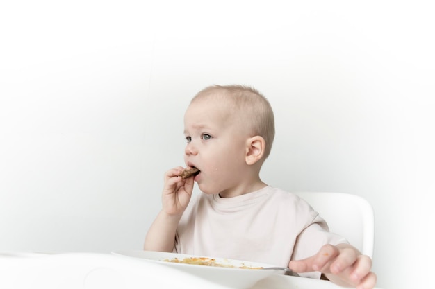 Un petit garçon mange de la soupe avec du pain tout seul dans une chaise haute sur fond de mur blanc