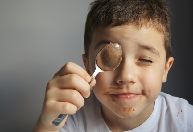 Photo petit garçon mangeant du chocolat à la cuillère. mur gris. mignon enfant heureux recouvert de chocolat