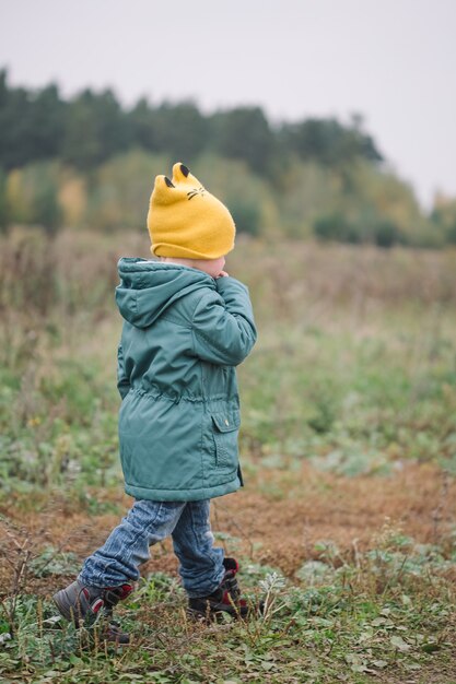 Petit garçon marchant dans le domaine dans un manteau vert et un chapeau jaune, vue latérale.