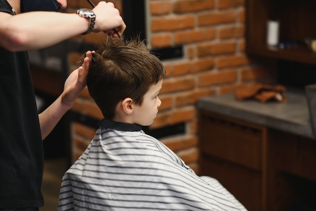 Un petit garçon mignon est assis chez un coiffeur chez le styliste, un écolier se fait couper les cheveux dans un salon de beauté, un enfant dans un salon de coiffure, une coupe courte pour homme.