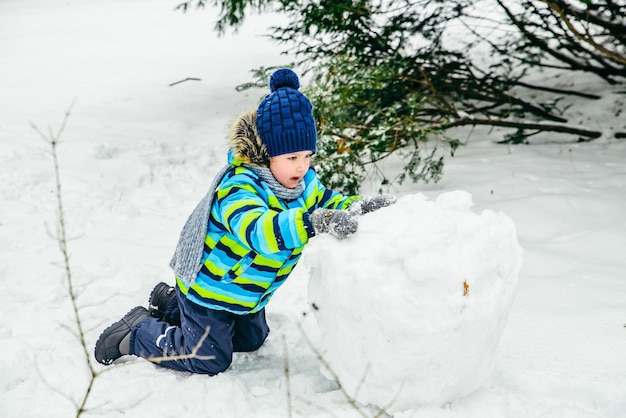 Petit garçon mignon faisant bonhomme de neige roulant grosse boule de neige