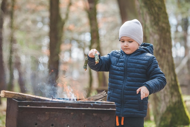 Un petit garçon a mis des branches au feu dans un grill reposant dans le concept de barbecue en forêt